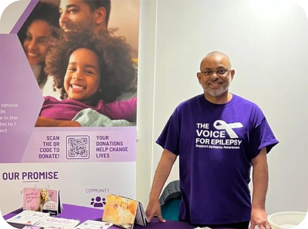 Smiling volunteer wearing The Voice For Epilepsy purple t-shirt at charity awareness stall with donation QR code and community materials