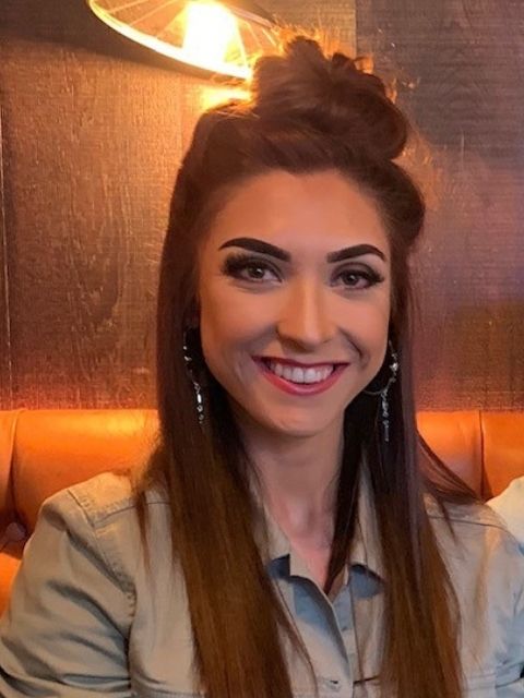 Woman with long brown hair styled in a bun, wearing earrings and a light denim shirt, smiling at the camera.