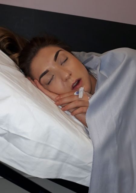 A young woman sleeping peacefully in a hospital bed, covered with a light blanket and connected to medical equipment.