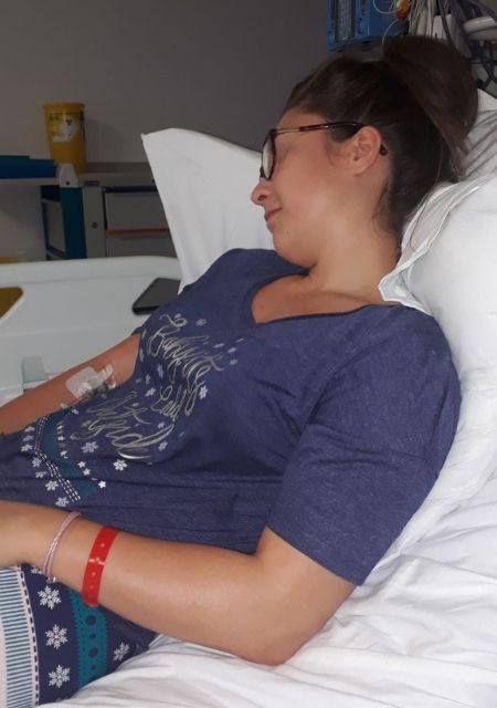 Woman resting in a hospital bed wearing a blue shirt with a medical IV line attached to her arm.