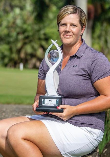 Female golfer holding a trophy while seated outdoors on a stone surface.