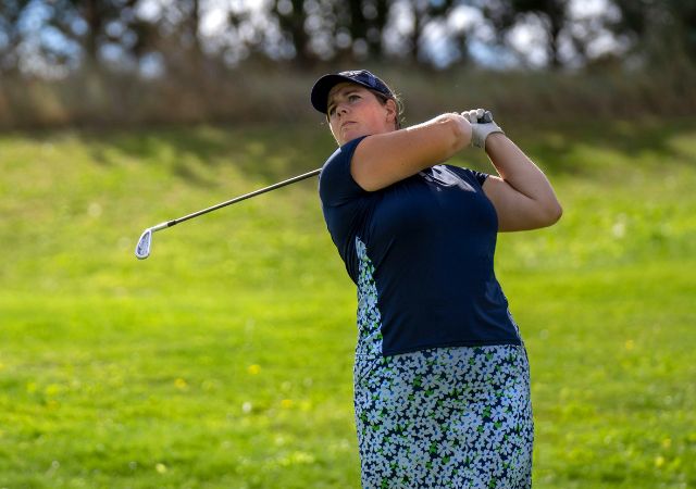 Female golfer in a blue top and floral skirt swinging a golf club on a green course.