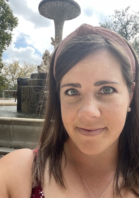 Woman with brown hair and a headband posing in front of a fountain in a park setting.