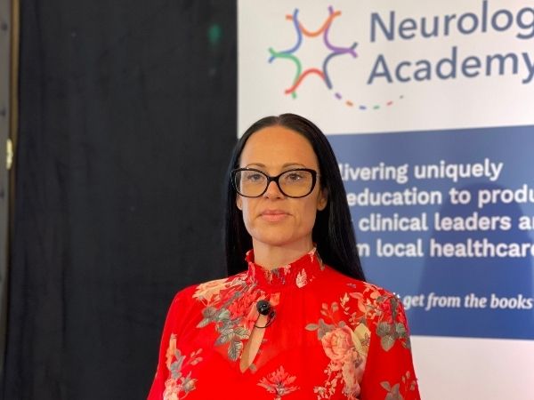 Woman wearing glasses and a red floral blouse standing in front of a Neurology Academy banner.